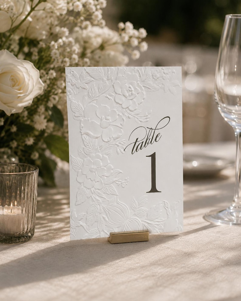 May include: A white rectangular table number card featuring an embossed floral pattern on the left side and the black printed text Table 1 on the right. The card is held upright by a small gold metal stand on a light-colored fabric surface.