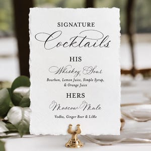 May include: A rectangular white card with deckled edges held by a gold metal stand. The text reads: SIGNATURE Cocktails, HIS Whiskey Sour, Bourbon, Lemon Juice, Simple Syrup, & Orange Juice, HERS Moscow Mule, Vodka, Ginger Beer & LiMe.