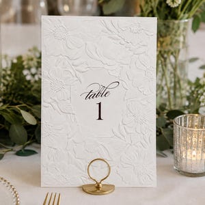 May include: A white rectangular table number card featuring an embossed floral pattern across the surface. The center of the card displays the text "table 1" in a black script and serif font. The card is held upright by a circular gold metal stand.