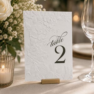 May include: A rectangular white table number card featuring an embossed floral pattern on the left side and the black printed text "Table 2" on the right. The card is held upright by a small, rectangular, metallic gold-colored stand.