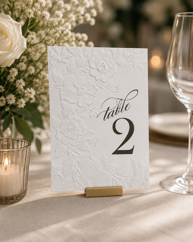 May include: A rectangular white table number card featuring an embossed floral pattern on the left side and the black printed text "Table 2" on the right. The card is held upright by a small, rectangular, metallic gold-colored stand.