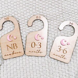 May include: Three wooden hangers with pink crescent moons and stars. The hangers are labeled "NB newborn", "0-3 months", and "3-6 months".