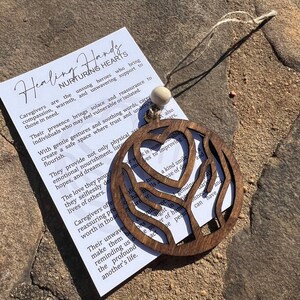 May include: Wooden ornament shaped like two hands holding a heart. The ornament is brown and has a natural wood finish. The text on the ornament reads "Healing Hands Nurturing Hearts".