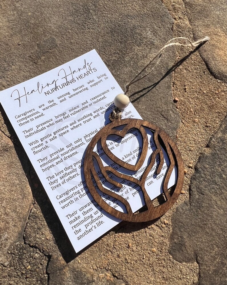 May include: Wooden ornament shaped like two hands holding a heart. The ornament is brown and has a natural wood finish. The text on the ornament reads "Healing Hands Nurturing Hearts".