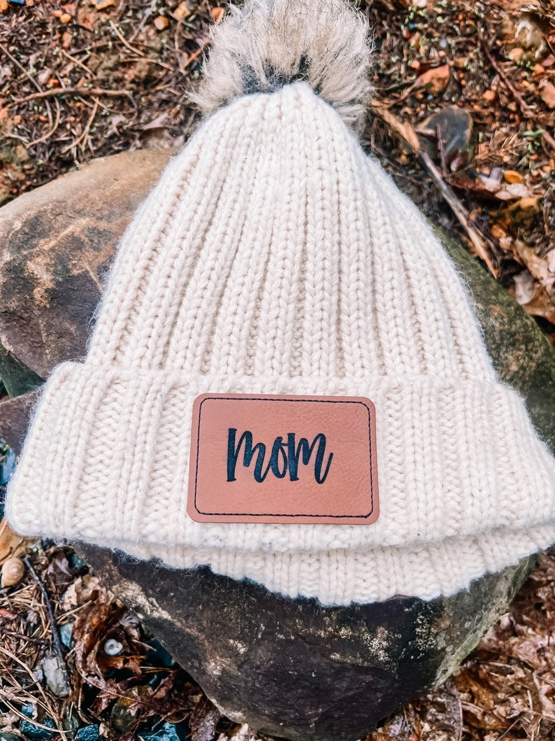 May include: Cream-colored knit beanie with a faux fur pom-pom on top. A brown leather patch with the word "mom" in black script is sewn on the front. The hat is made of a thick, ribbed knit material.