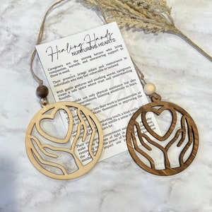 May include: Two wooden ornaments featuring a heart and hands design. The ornaments are brown and natural wood. The text on the card reads "Healing Hands Nurturing Hearts". Caregivers are the unsung heroes who bring compassion, warmth, and unwavering support to those in need.