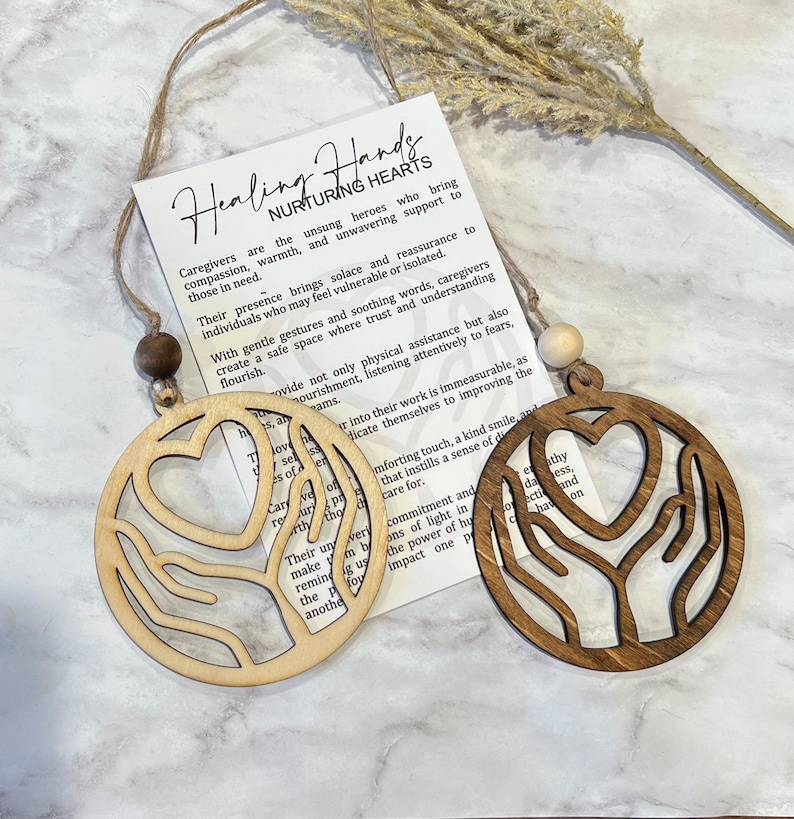May include: Two wooden ornaments featuring a heart and hands design. The ornaments are brown and natural wood. The text on the card reads "Healing Hands Nurturing Hearts". Caregivers are the unsung heroes who bring compassion, warmth, and unwavering support to those in need.