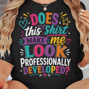 May include: A black short-sleeve t-shirt featuring colorful graphic text that reads: DOES this Shirt MAKE ME LOOK PROFESSIONALLY DEVELOPED? The design includes decorative elements like hearts, stars, and lines in shades of pink, blue, orange, and purple.