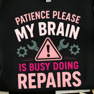 May include: Black crew neck sweatshirt with the text "PATIENCE PLEASE MY BRAIN IS BUSY DOING REPAIRS" in white and pink. The design includes wrenches, gears, and a caution sign. A casual, comfortable top.