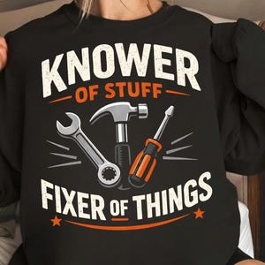 Puede incluir: Sudadera negra con el texto "KNOWer OF STUFF FIXER OF THINGS" en blanco y naranja. El diseño incluye ilustraciones de una llave inglesa, un martillo y un destornillador.