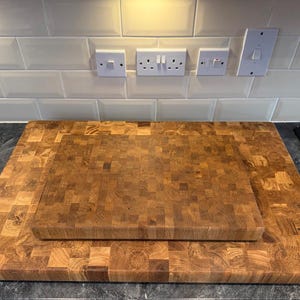 May include: Two wooden butcher block cutting boards. The larger board is rectangular, and the smaller board is placed on top. Both boards have a mosaic pattern of wood pieces in shades of brown. The boards are on a dark countertop.