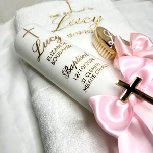 Pode incluir: Uma vela branca com uma cruz dourada e o nome "Lucy Elizabeth Boudaher" e o texto "Batizada 12/10/2024 St Clement Melkite Church" em letras douradas. Uma toalha branca com o nome "Lucy" bordado em letras douradas. Uma escova de cabelo dourada com uma letra "L" dourada. Uma fita rosa com uma cruz dourada.