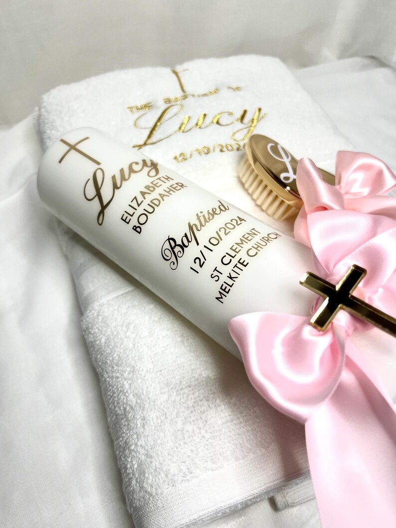 Pode incluir: Uma vela branca com uma cruz dourada e o nome "Lucy Elizabeth Boudaher" e o texto "Batizada 12/10/2024 St Clement Melkite Church" em letras douradas. Uma toalha branca com o nome "Lucy" bordado em letras douradas. Uma escova de cabelo dourada com uma letra "L" dourada. Uma fita rosa com uma cruz dourada.
