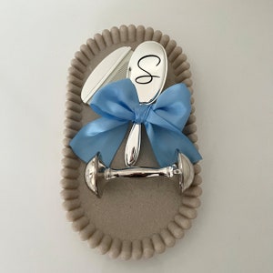 Puede incluir: Un juego de regalo para bebé sobre una bandeja ovalada decorativa. El juego incluye una cuchara plateada con la letra "S", un peine y un sonajero, todo atado con una cinta azul claro. La bandeja es beige con un borde con cuentas.