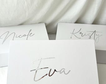 Puede incluir: Tres cajas de regalo blancas con letras plateadas. Las cajas están etiquetadas con "Nicole", "Kristy" y "Eva" en una fuente cursiva. Las cajas están sobre un fondo de tela blanca.