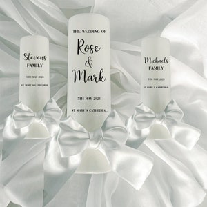 Puede incluir: Dos velas de unidad blancas con lazos de satén blanco. Las velas están decoradas con texto negro que dice "The Wedding of Rose & Mark" y "5th May 2023" y "St Mary's Cathedral". La otra vela dice "Stevens Family" y "Michaels Family".