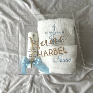 Könnte beinhalten: Eine transparente Acrylbox gefüllt mit einem weißen Handtuch, einer weißen Kerze mit goldenem Kreuz und einem blauen Band. Das Handtuch ist mit dem Text "Die Taufe von Isaac 2023" und "Isaac" bestickt. Die Kerze trägt den Text "Isaac Harbel" und ein Kreuz.