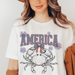 May include: White t-shirt with a red, white, and blue crab design. The crab is wearing a bow and the word "AMERICA" is written above it. There are fireworks around the crab.