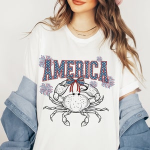 May include: White t-shirt with a red, white, and blue crab design. The crab is wearing a red, white, and blue bow. The text "AMERICA" is written in blue with white stars above and below the letters. There are red, white, and blue fireworks on either side of the crab.