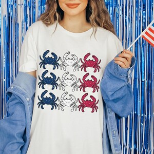 May include: A white t-shirt with a pattern of red, white, and blue crabs. The shirt is being worn by a person holding a small American flag.