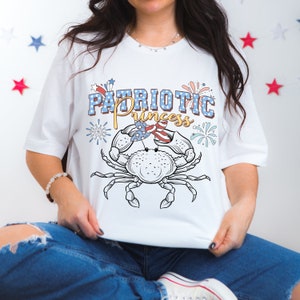 May include: White t-shirt with a red, white, and blue crab wearing a headband with the American flag. The text "PATRIOTIC Princess" is above the crab.