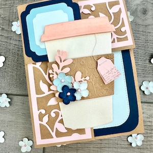 May include: Handmade greeting card featuring a coffee cup design with floral embellishments. The card has a light pink lid, a white cup, and a brown sleeve with embossed details. Decorative paper flowers and a tea bag tag add charm.
