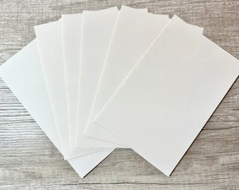 May include: A stack of six white blank cards, arranged in a fan shape on a light wood surface.