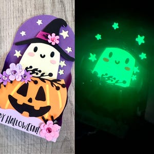 May include: A layered paper craft card featuring a white ghost wearing a black witch hat, a bright orange jack-o'-lantern, and purple flowers. The design includes small stars and the text "HAPPY HALLOWEEN!" with the ghost and stars glowing green in the dark.