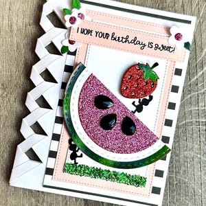 May include: A handmade greeting card featuring a pink glittery watermelon slice with black teardrop-shaped seeds, a red glittery strawberry, and white flowers with pink centers. The text reads I HOPE YOUR birthday IS SWEET! on a light pink banner.
