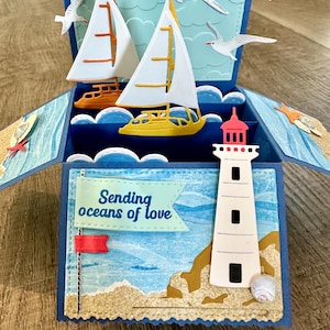 May include: A three-dimensional pop-up greeting card featuring a nautical theme. The card displays two sailboats, white seagulls, and a lighthouse. A light blue banner on the front includes the text Sending oceans of love in dark blue lettering.