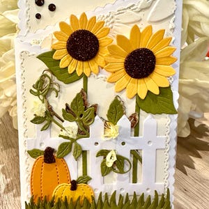 May include: A handmade greeting card featuring two yellow sunflowers with dark brown glitter centers, two orange pumpkins, and a white picket fence. The card has a scalloped edge, green die-cut grass, and embossed cream-colored cardstock layers.