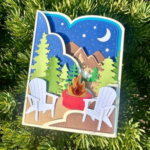 May include: A layered paper craft greeting card featuring a night scene with a crescent moon, stars, brown mountains, green pine trees, a red brick fire pit with orange flames, and two white chairs arranged on a brown ground surface.