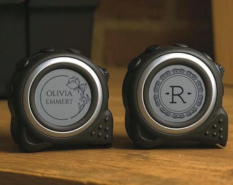 May include: Two black tape measures with silver accents and personalized details. One features the name "OLIVIA EMMERT" within a floral design, the other displays the letter "R" in a decorative frame. They sit on a wooden surface.