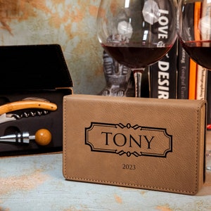 May include: A brown leatherette box with the name "TONY" engraved on the front. The box contains a wine opener, a bottle stopper, and a corkscrew. The box is sitting on a wooden table next to a bottle of wine and two wine glasses.