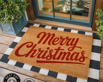 May include: A rectangular doormat with the words "Merry Christmas" in red script. The mat is tan with a black and white checkered border. The mat is placed in front of a door.