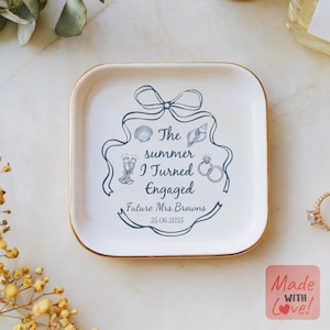 May include: A square, white ceramic ring dish with a gold rim. The dish features a dark blue design with the text "The summer I Turned Engaged" and the date "25.06.2025". Decorative elements include seashells, champagne flutes, and a diamond ring.