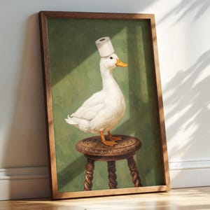 May include: A framed wall art print featuring a white duck with an orange beak standing on a round, carved wooden stool. The duck has a roll of white toilet paper balanced on its head. The background is a solid, muted olive green color.