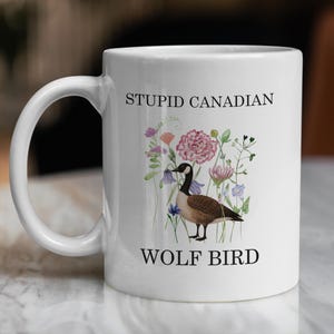 May include: White ceramic mug with the text "STUPID CANADIAN" and "WOLF BIRD". The mug features a watercolor illustration of a Canada goose among colorful flowers. The handle is white.