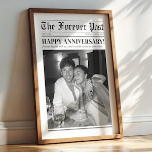 May include: A framed black and white photograph resembling a newspaper front page. The headline reads "HAPPY ANNIVERSARY!" with the text "The Forever Post" at the top. The image features a couple embracing, with a wooden frame.