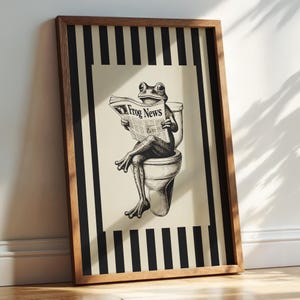 May include: Framed art print featuring a frog reading a newspaper titled "Frog News" while seated on a toilet. The artwork is in black and white, set against a cream background with a black and white striped border, and framed in wood.