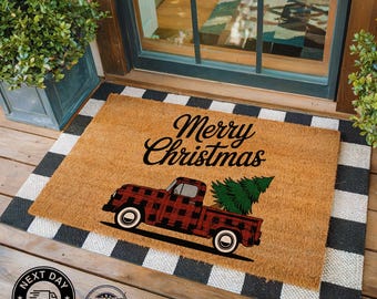 May include: A rectangular doormat with a black and white checkered border. The center features a brown background with a red and black plaid truck carrying a green Christmas tree. The words "Merry Christmas" are written in black script.