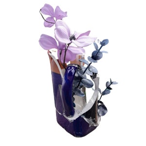 May include: A broken blue and white ceramic vase with a purple and white flower arrangement. The vase is decorated with a blue and white pattern. The flowers are arranged in a cluster and are mostly purple with some white flowers.
