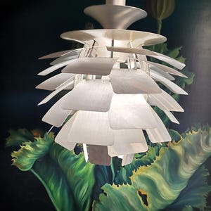 Peut inclure: Un luminaire suspendu moderne argenté avec un design en couches en forme de cœur d'artichaut. Le luminaire est suspendu à un support de plafond et comporte plusieurs panneaux rectangulaires superposés. Le fond montre un mur sombre avec une peinture florale verte.
