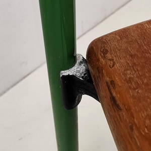 May include: Close-up of a green metal chair leg with a black metal bracket connecting to a wooden seat. The wood has a natural grain pattern.