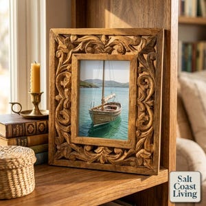 May include: A square wooden picture frame featuring intricate hand-carved floral scrollwork patterns along the border. The frame holds a photograph of a wooden sailboat on calm blue water, set against a backdrop of distant green hills.