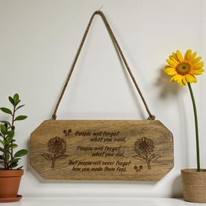 May include: Wooden sign with a brown rope for hanging. The sign has engraved text and sunflower illustrations. The text reads: "People will forget what you said, People will forget what you did, But people will never forget how you made them feel."