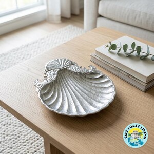 May include: A silver-toned metal decorative dish shaped like a scallop shell. The dish features a textured, ribbed surface radiating from the center to the scalloped edges, with detailed, ornate relief carvings at the top hinge area of the shell.