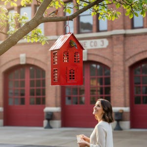 May include: A bright red, house-shaped bird feeder hanging from a tree branch. The feeder has multiple windows and a door, with a small bird perched on top. The background features a brick building with red doors and the text "FIRE CO 3".