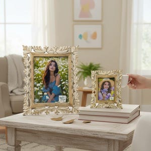 May include: Two decorative, off-white picture frames with floral and leaf carvings. The larger frame holds a photo of a woman in a blue dress, while the smaller frame displays a woman in a hat. Both frames are on a light-colored wooden table.