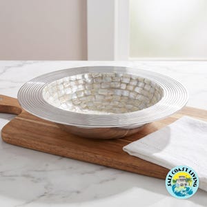 May include: A round decorative bowl featuring a wide, silver-toned metallic rim with concentric circular grooves. The interior of the bowl is lined with a mosaic of small, iridescent white mother-of-pearl tiles arranged in a grid pattern.